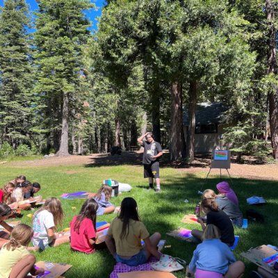 Children sitting on the grass outside, surrounded by trees, painting colorful artwork.